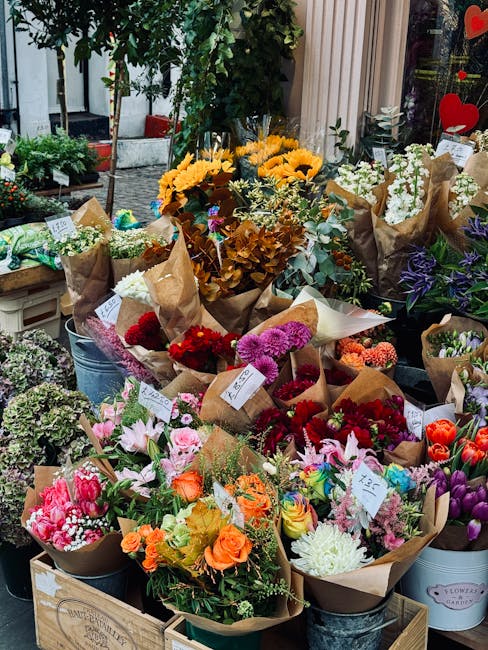 A vibrant display of fresh flower bouquets arranged outdoors on a wooden table, featuring a variety of colorful blooms including sunflowers, roses, lilies, tulips, and chrysanthemums, wrapped in brown