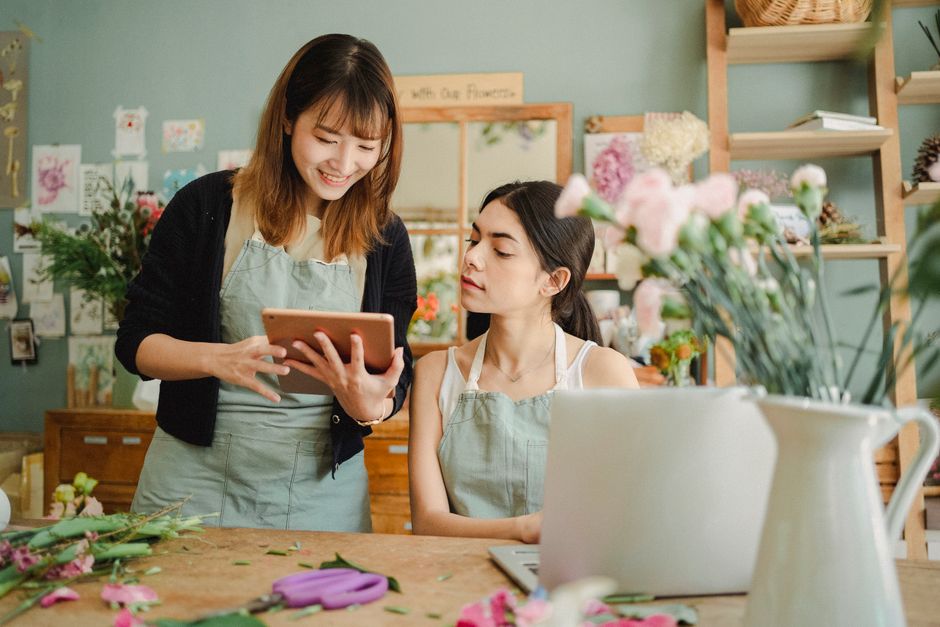 Two women in a floral workshop or flower shop, with a wooden work table displaying various flower arrangements and materials. The woman on the left, with shoulder-length brown hair, is smiling and hol