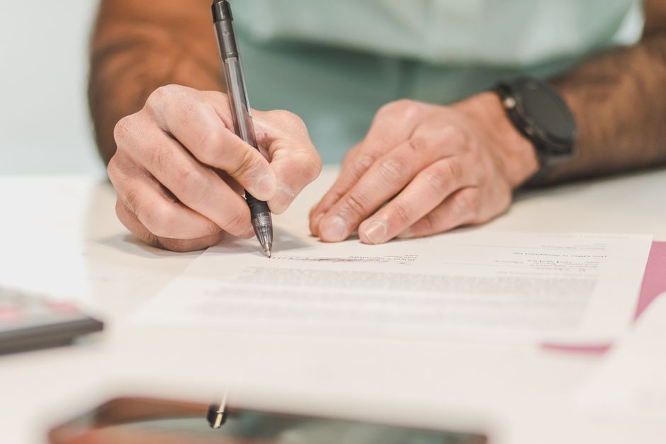A person with a silver pen is signing a document on a white table, with their left hand resting nearby. The individual wears a black watch on their right wrist, and the background is softly blurred, f