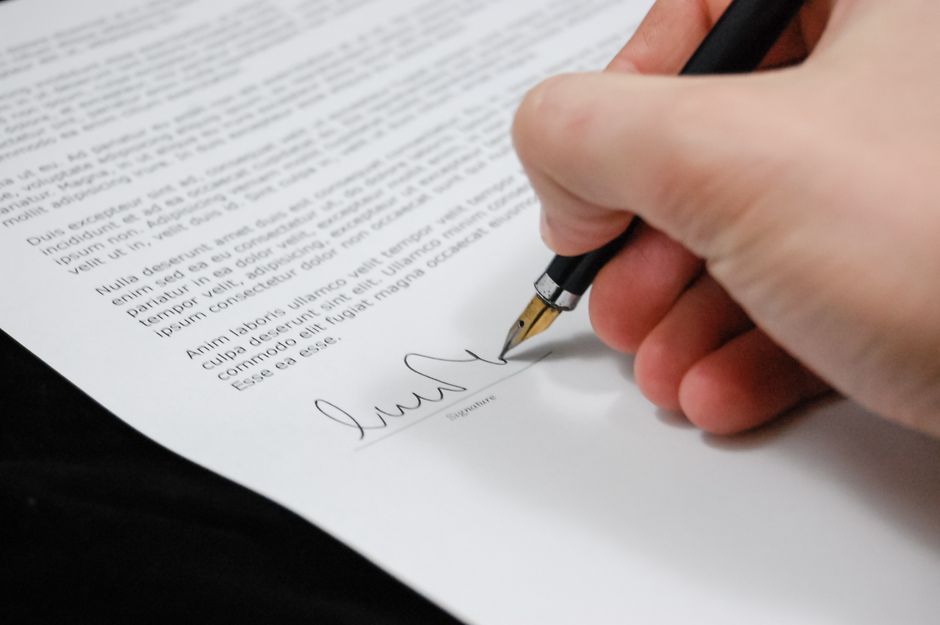 Close-up of a hand holding a black and gold fountain pen, signing a document with printed text. The hand appears to be mature, with neatly trimmed nails. The paper is white, placed on a dark surface, 
