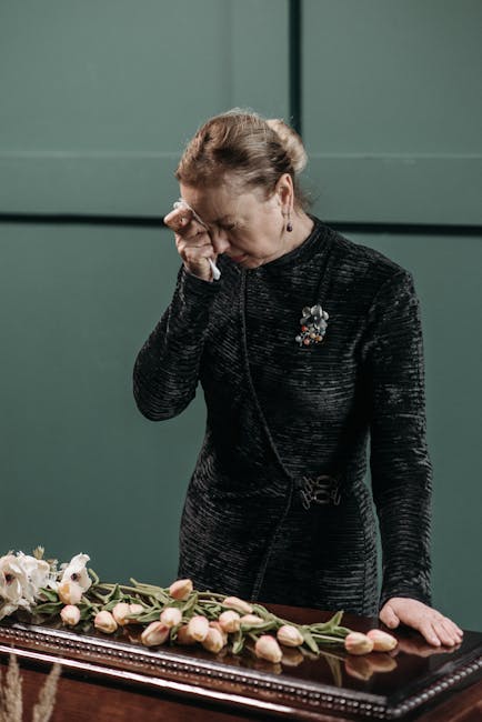 A woman dressed in a black textured dress with a floral brooch is standing beside a table, visibly emotional and wiping her eye with her right hand. On the table in front of her, there is a bouquet ar