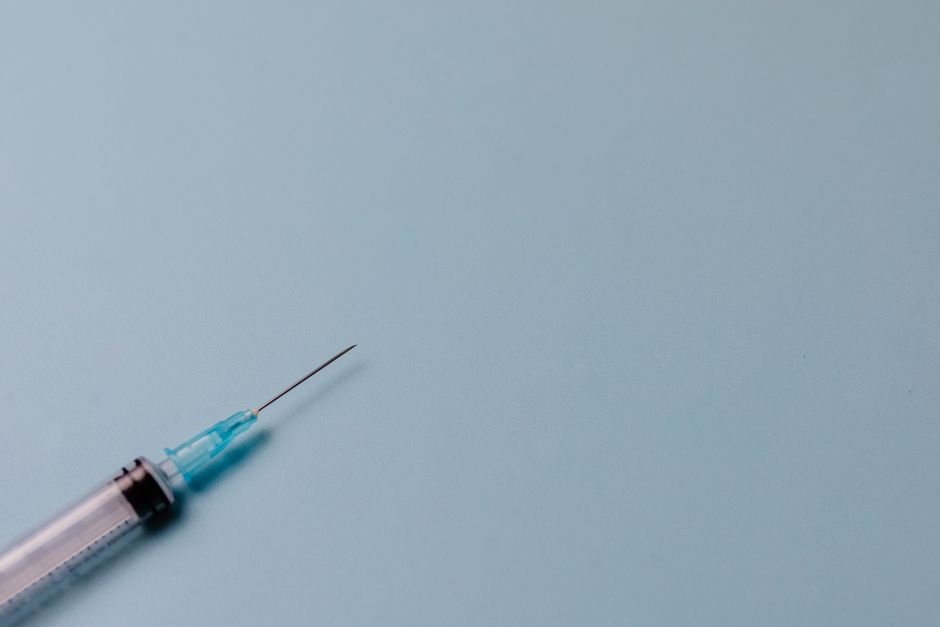 A close-up view of a medical syringe with a thin, sterile needle and a transparent barrel filled with clear liquid, resting on a smooth, light blue surface. The syringe is positioned diagonally across