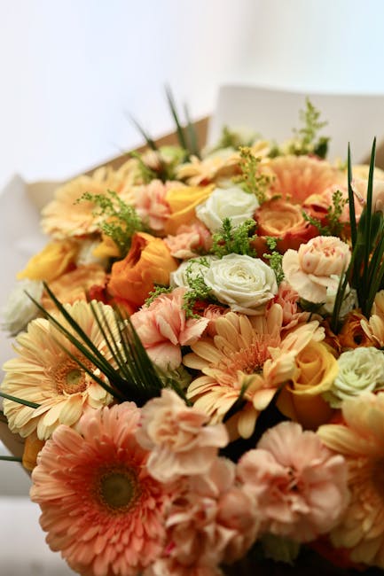 A vibrant bouquet of fresh flowers showcasing a mix of peach, cream, and soft pink gerbera daisies, roses, and eustoma, accented with green foliage including long palm leaves and delicate sprigs. The 